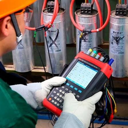 Technician holding a three-phase power quality analyzer, test leads on an outdoor capacitor bank.