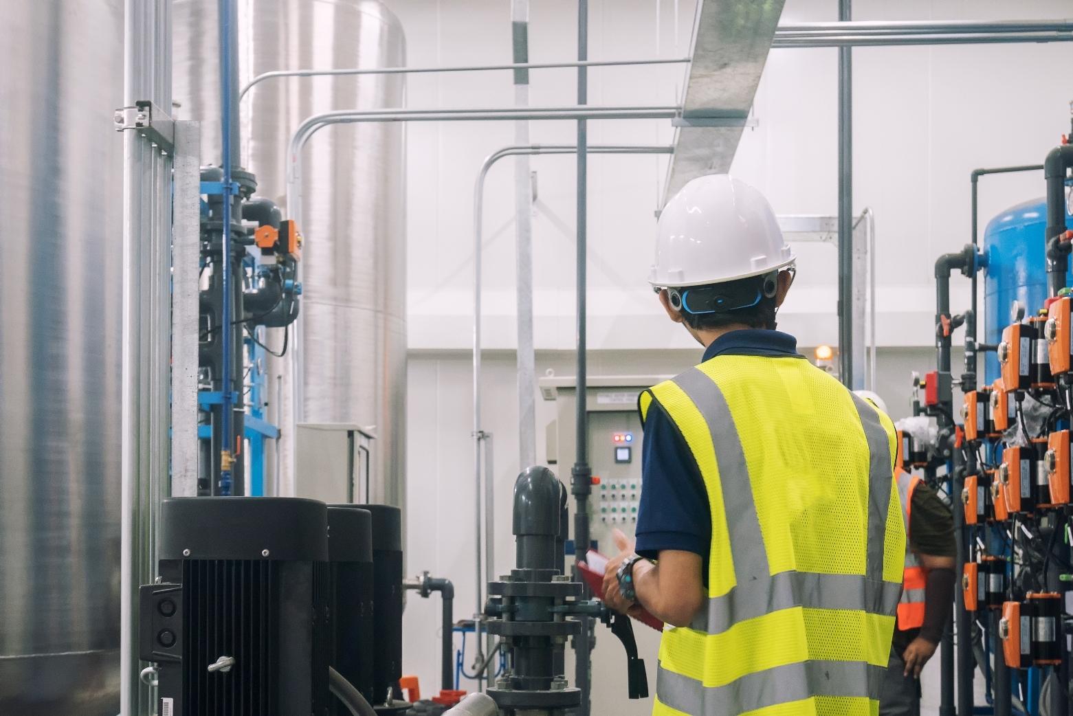 Maintenance technician in a hi-vis vest and hard hat inspecting equipment panels in a plant.