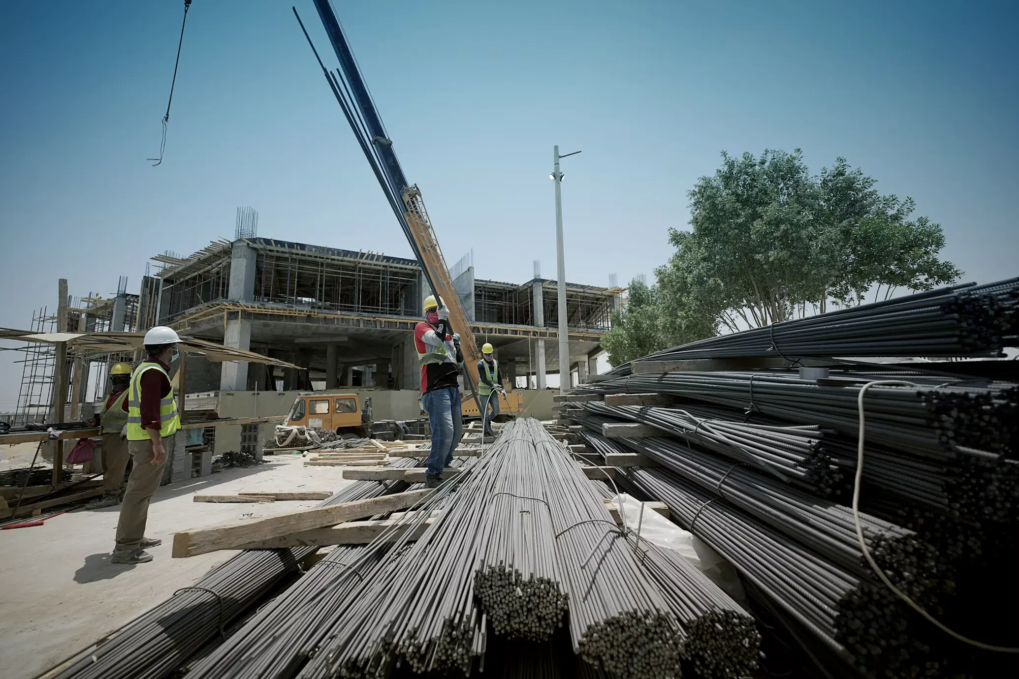 Sitio en construcción con trabajadores en equipo reflectante, una grúa y varilla apilada.